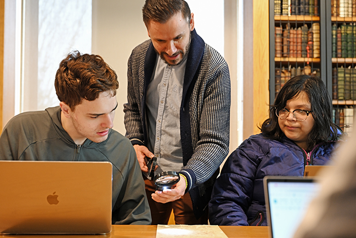 Klein Hernández uses a magnifying class to highlight the details of a historical document for “Borders, Empire, Immigration” students Christian Pappas ’26, left, and Danna Sandoval ’24.